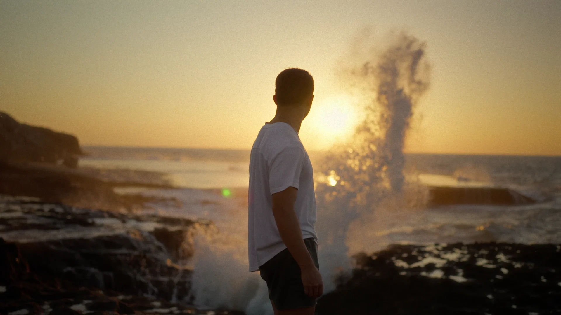A figure at the shoreline as a wave rises in the last light.